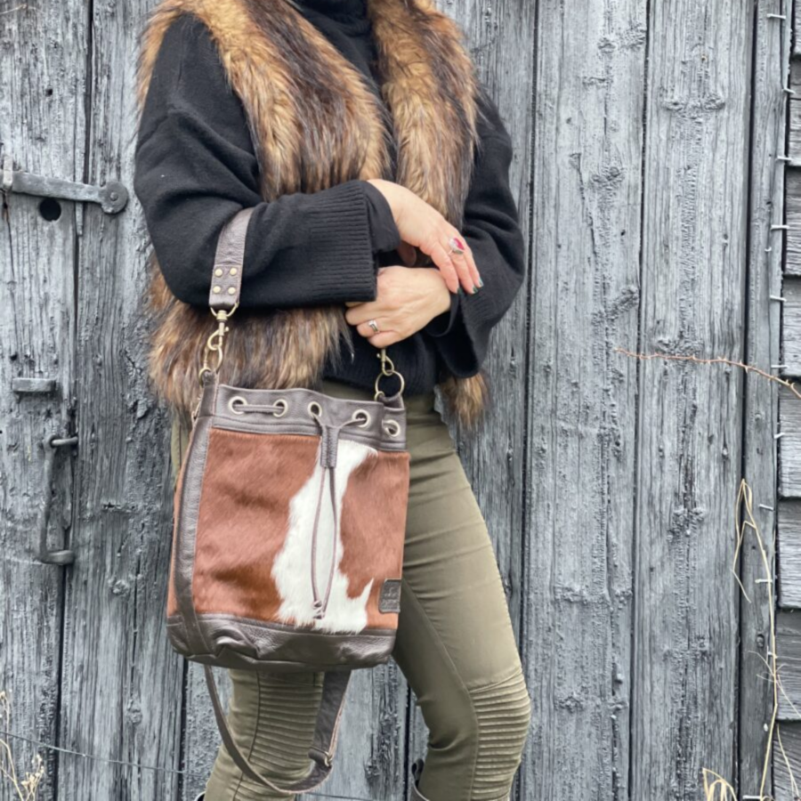 Woman leaning against a wall, wearing a brown and white cowhide bucket bag, showcasing the bag's relaxed style
